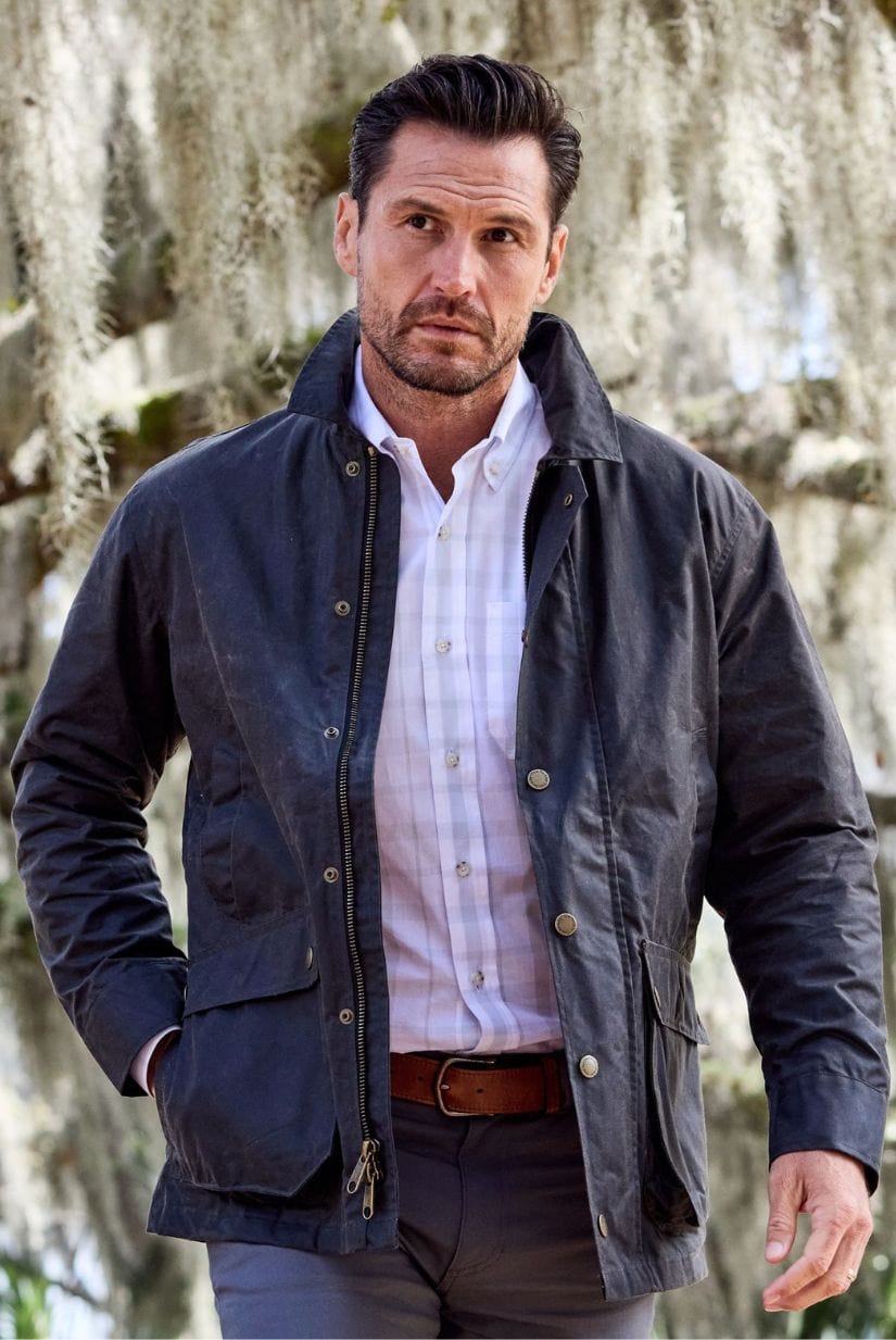 A man in a dark jacket and white shirt stands outdoors with moss-covered trees in the background.