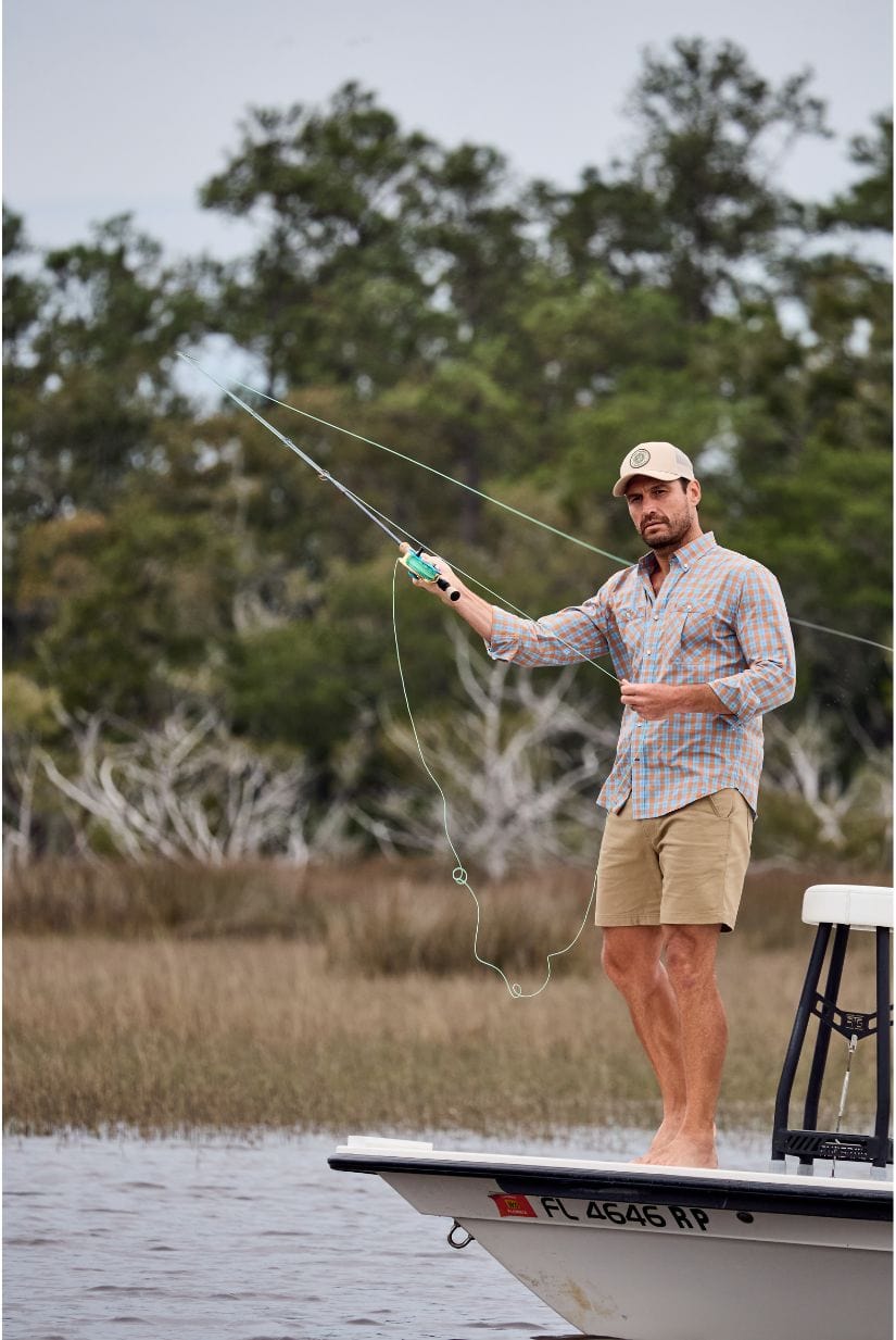 Man in a plaid shirt and shorts fly fishing barefoot from a small boat in a marshy area, surrounded by trees.