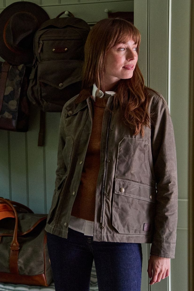 Woman in a brown jacket and sweater stands indoors near bags and coats, looking to the side.