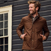 Man stands outside of a house with hands in front pockets of a brown quilted jacket with collar.