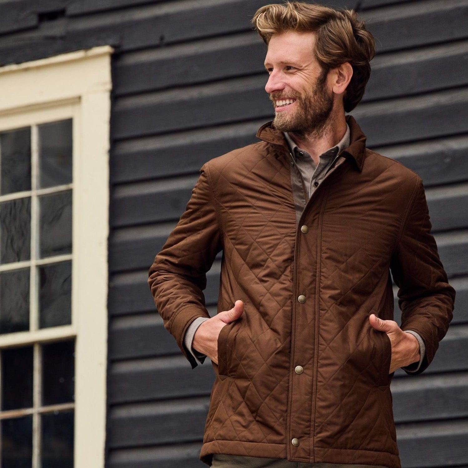 Man stands outside of a house with hands in front pockets of a brown quilted jacket with collar.