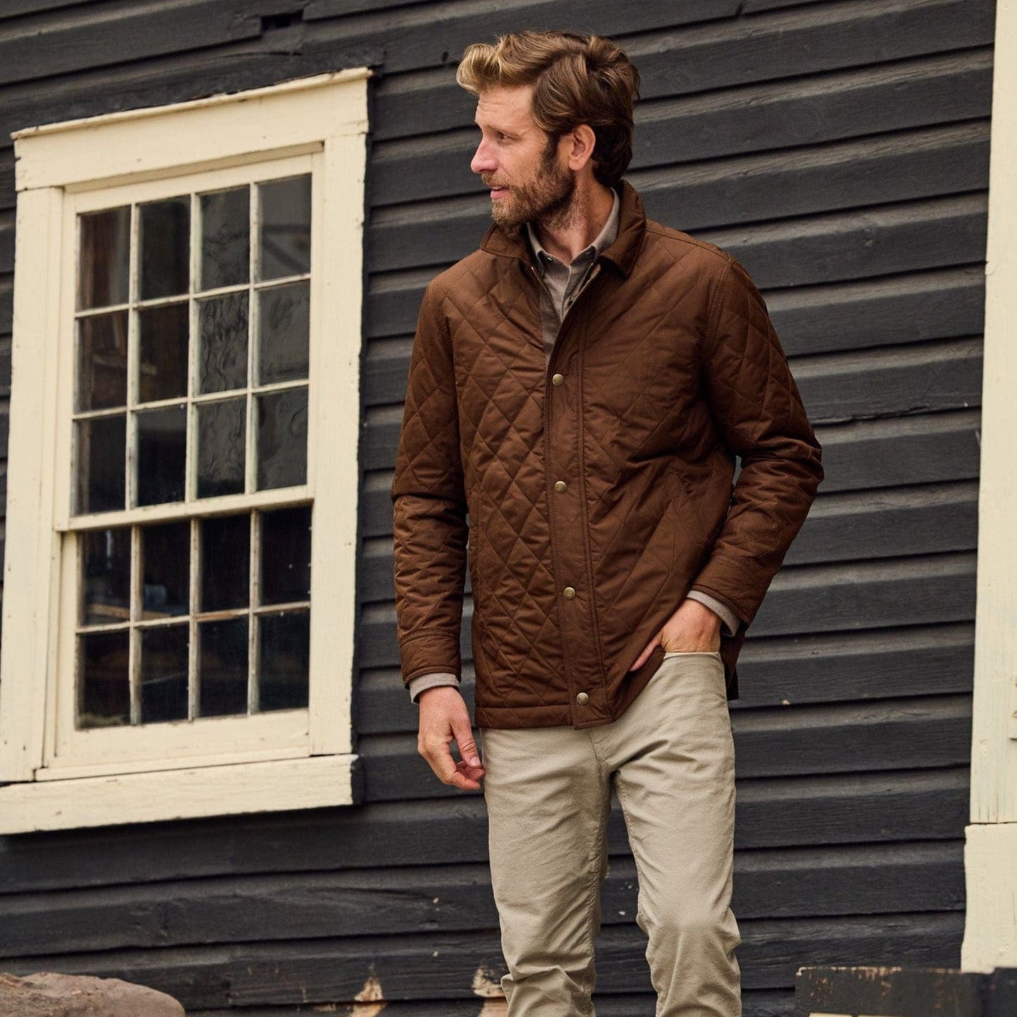 Man stands outside of a house wearing a brown quilted jacket with collar and khaki redmont pants.