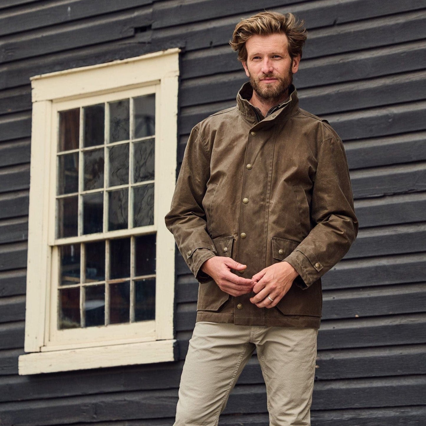 A man stands before a wooden building in a Tom Beckbe Tensaw Jacket and beige pants.