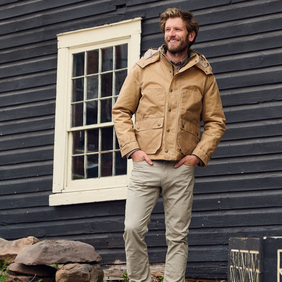 A man in a weatherproof Fowler Jacket by Tom Beckbe and beige pants smiles in front of a dark wooden building with a window.