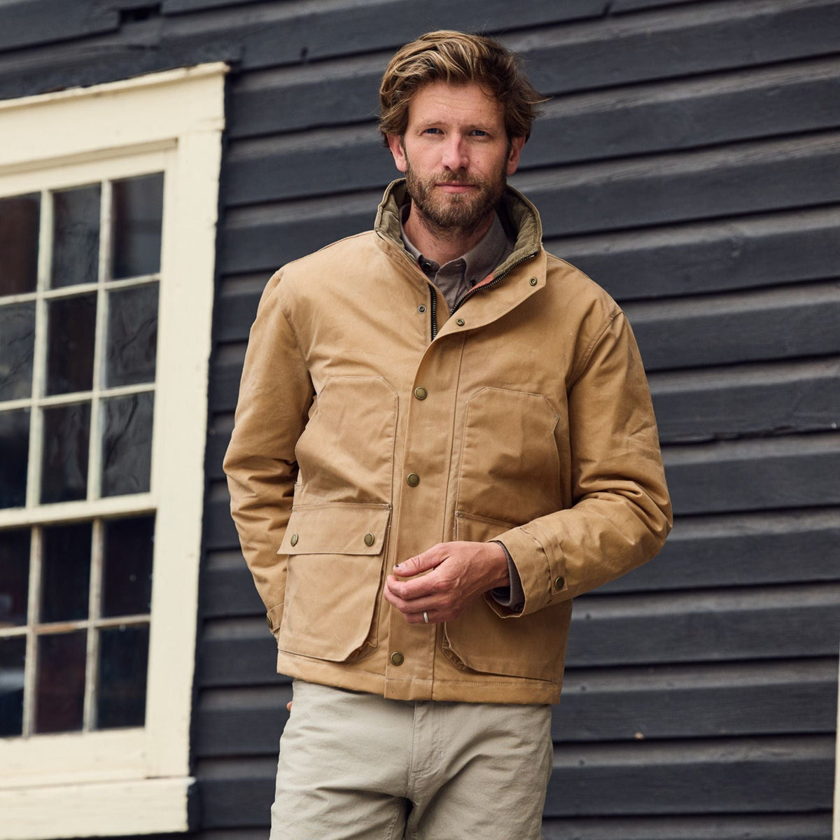 A bearded man stands outside in a Tom Beckbe Fowler Jacket and khaki pants by a dark wooden wall with a window.