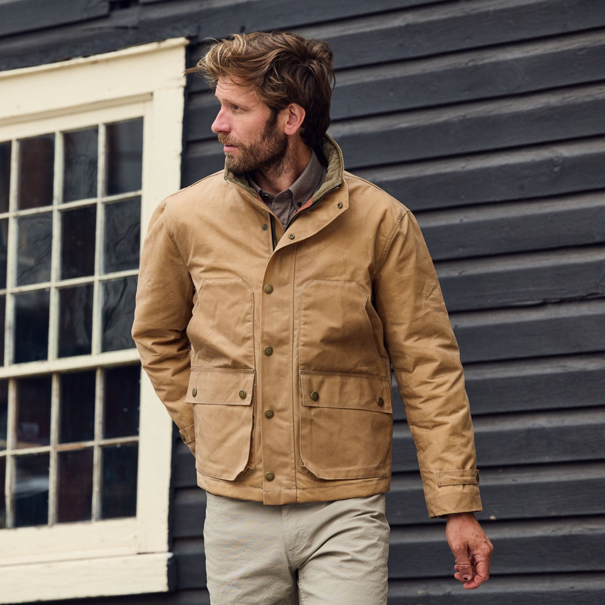 A man in a Tom Beckbe Fowler Jacket and light pants stands before a dark wooden building, looking to his left.