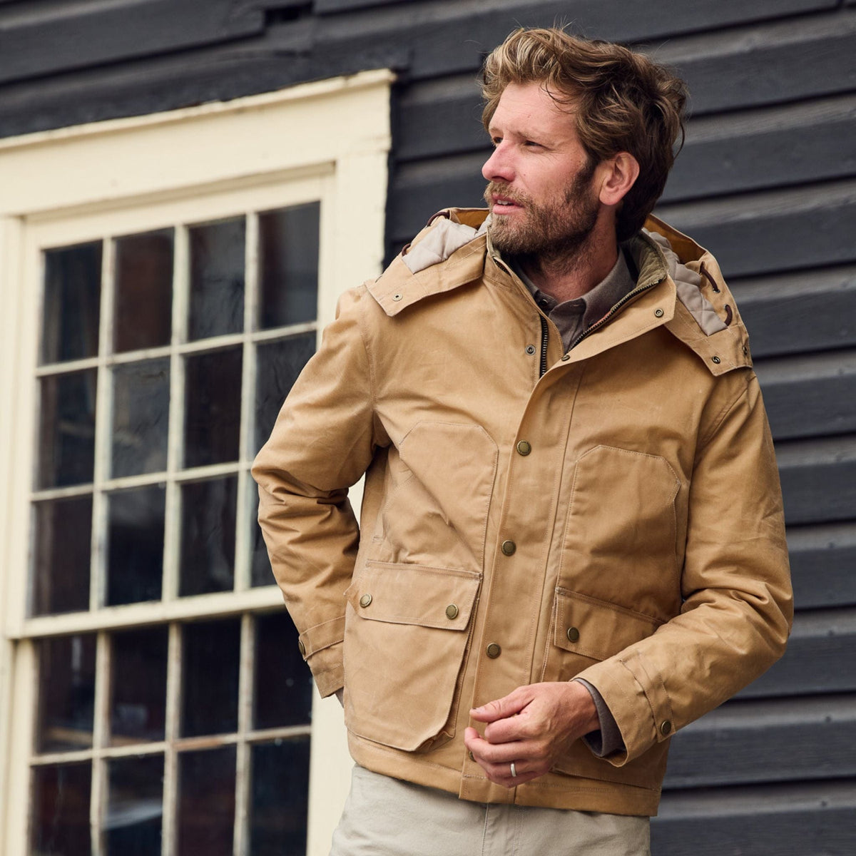 A man in a Tom Beckbe Fowler Jacket stands before a black wooden building with white-framed windows, looking to the side.