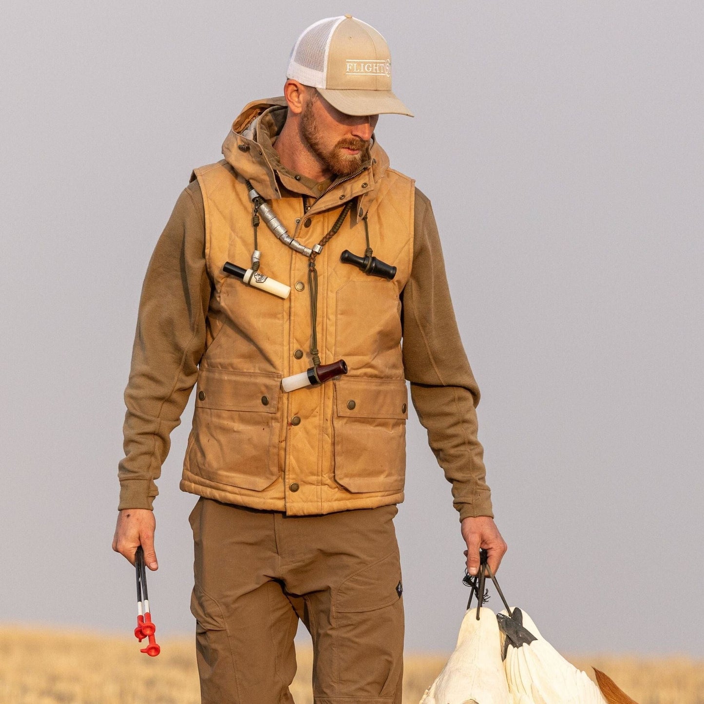 A man in a Tom Beckbe Fowler Vest and beige cap holds a plastic clamp and carry strap, standing in a field.