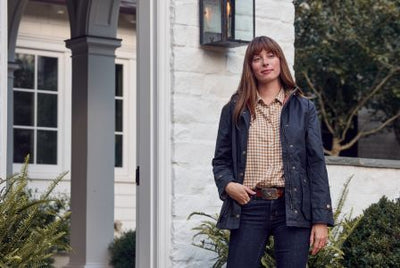 Woman standing outside a white brick house, wearing a dark jacket, checked shirt, and jeans, surrounded by greenery.