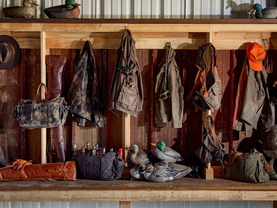 Row of outdoor gear: hunting jackets, backpacks, hats, and decoys on wooden shelves; assorted bags and boots below.