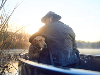 A man in a hat sits in a canoe on a calm lake, holding a dog. The sun is setting, casting a warm glow over the scene.