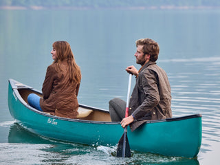 Two people in brown jackets canoeing on a calm lake. The woman looks ahead while the man paddles at the rear.