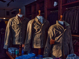 Three brown jackets on mannequins in a clothing store, with folded shirts displayed below on a shelf.