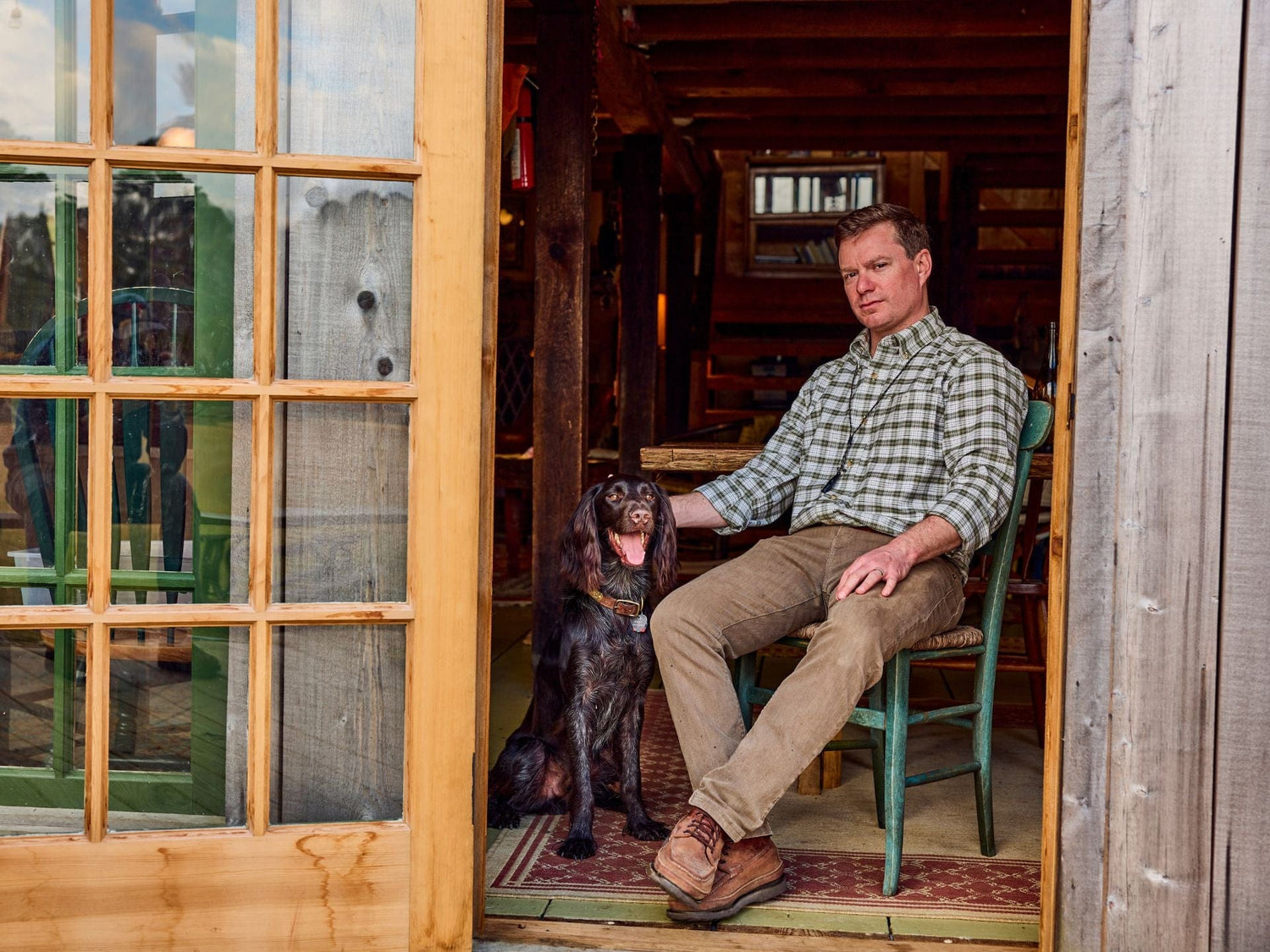 Man sitting on a chair in a cabin next to a dog, with a wooden door partially open.