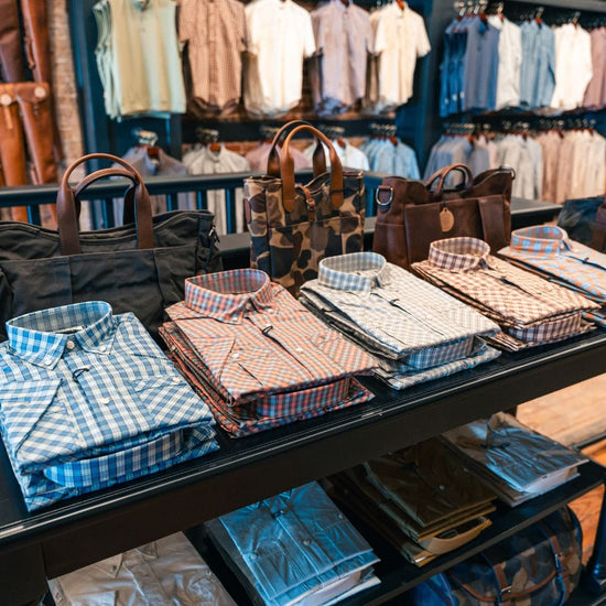 Folded plaid shirts and bags displayed on a store shelf with a variety of shirts hanging in the background.