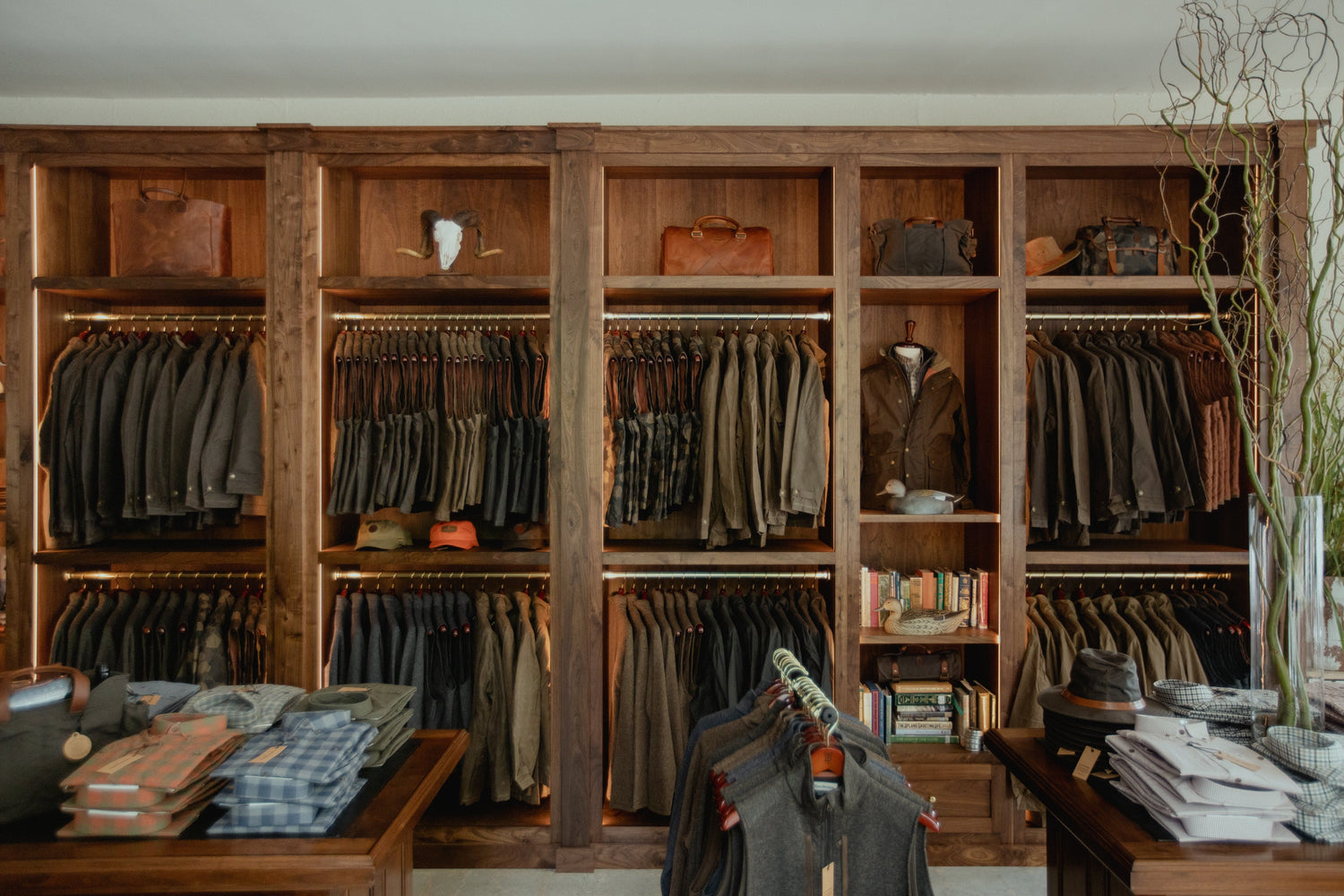 Wooden shelves filled with neatly arranged mens clothing, hats, and bags. Tables display folded shirts and hats in the foreground.