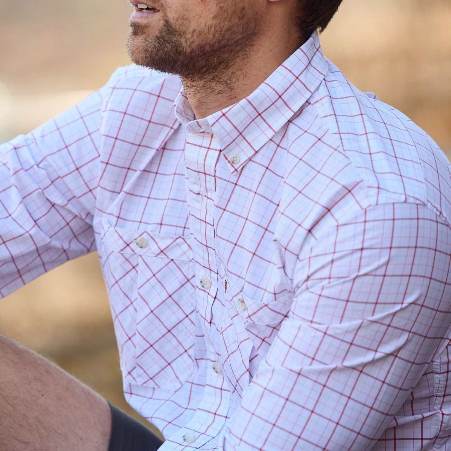 A person in a Tom Beckbe Tidewater Shirt with plaid design and rolled sleeves sits outdoors against a blurred natural backdrop.