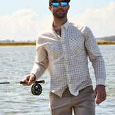Person in water holding a rod, wearing a Tom Beckbe Tidewater Shirt and cap, against sky and water.