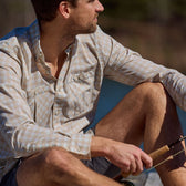 Man in a Tom Beckbe Tidewater Shirt and shorts sits by the water with a fishing rod, looking away from the camera.