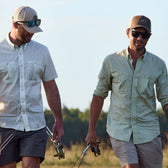 Two men in Tidewater shirts by Tom Beckbe with UPF 30+ walk on grass holding fishing rods.