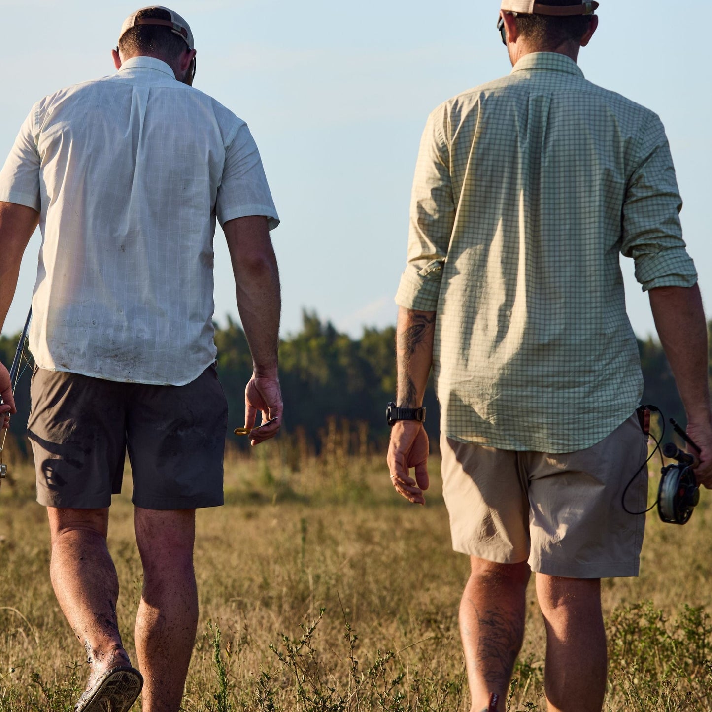 Two men in Tom Beckbe Tidewater Shirts stroll through a grassy field, rods in hand, under a clear sky.
