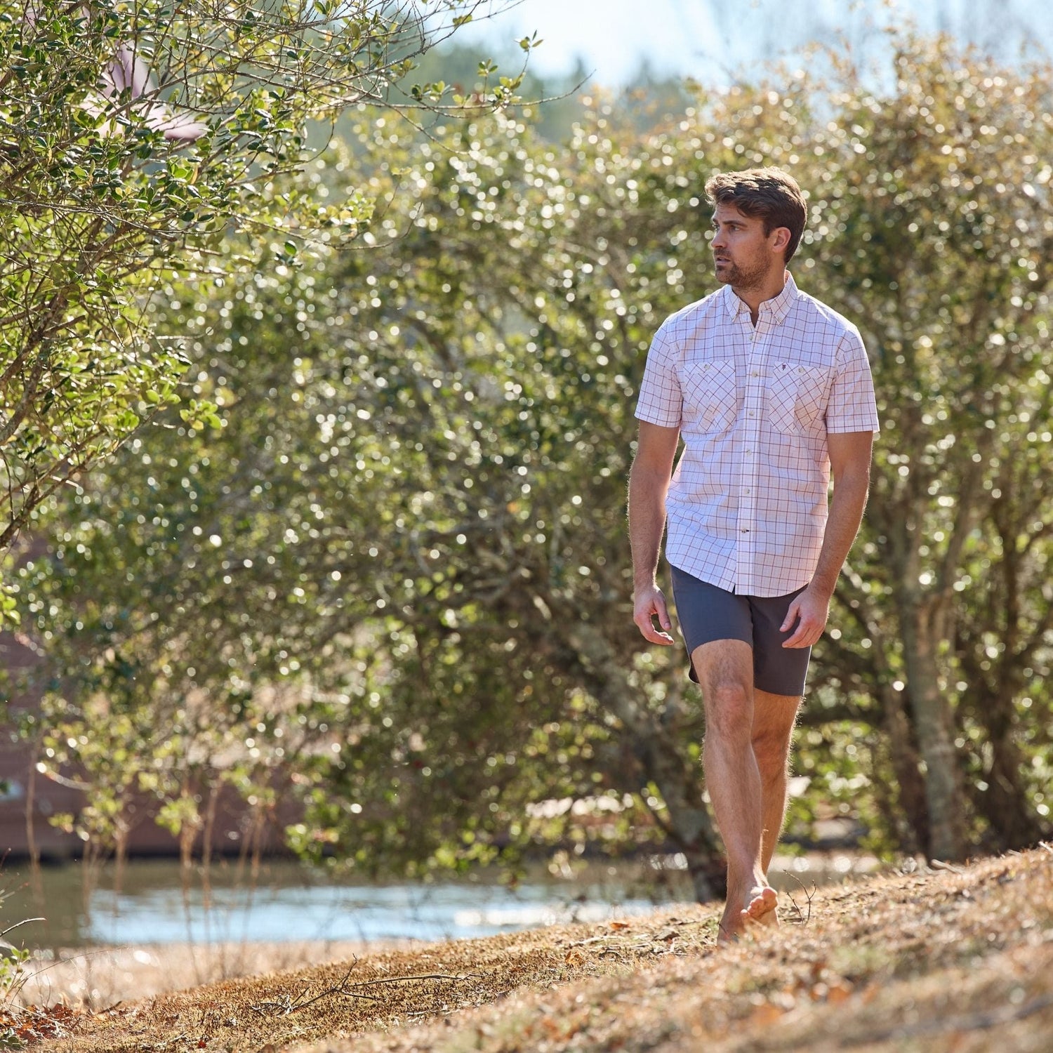 A man in a Tom Beckbe Tidewater Shirt and shorts strolls barefoot on grass near water, with trees in the background.