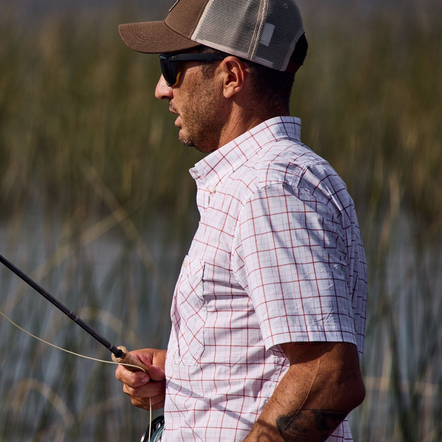 A man in a Tom Beckbe Tidewater Shirt, wearing sunglasses, enjoys fishing with UPF 30+ coverage near tall grass.