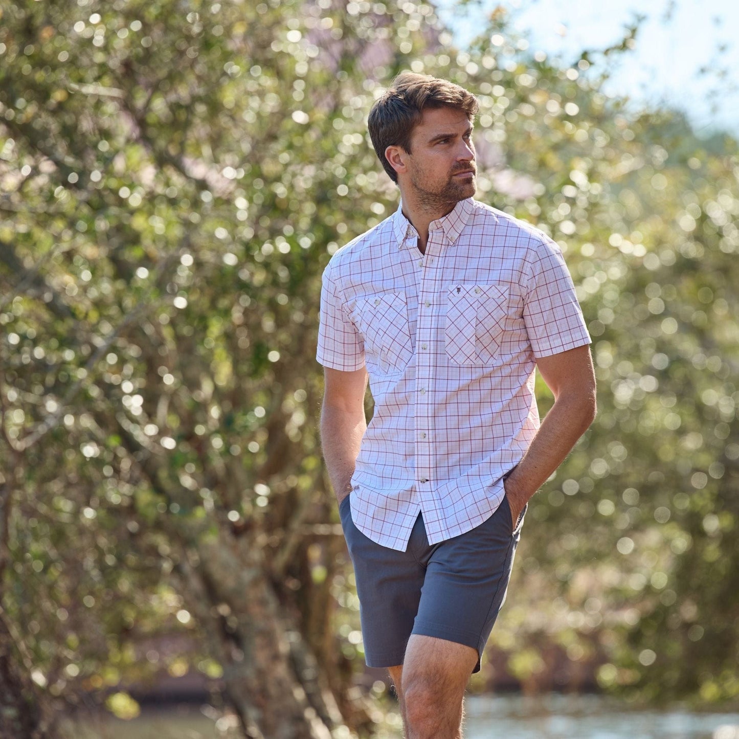 Man wearing a Tidewater Shirt by Tom Beckbe and shorts, strolls outside with trees in the background.