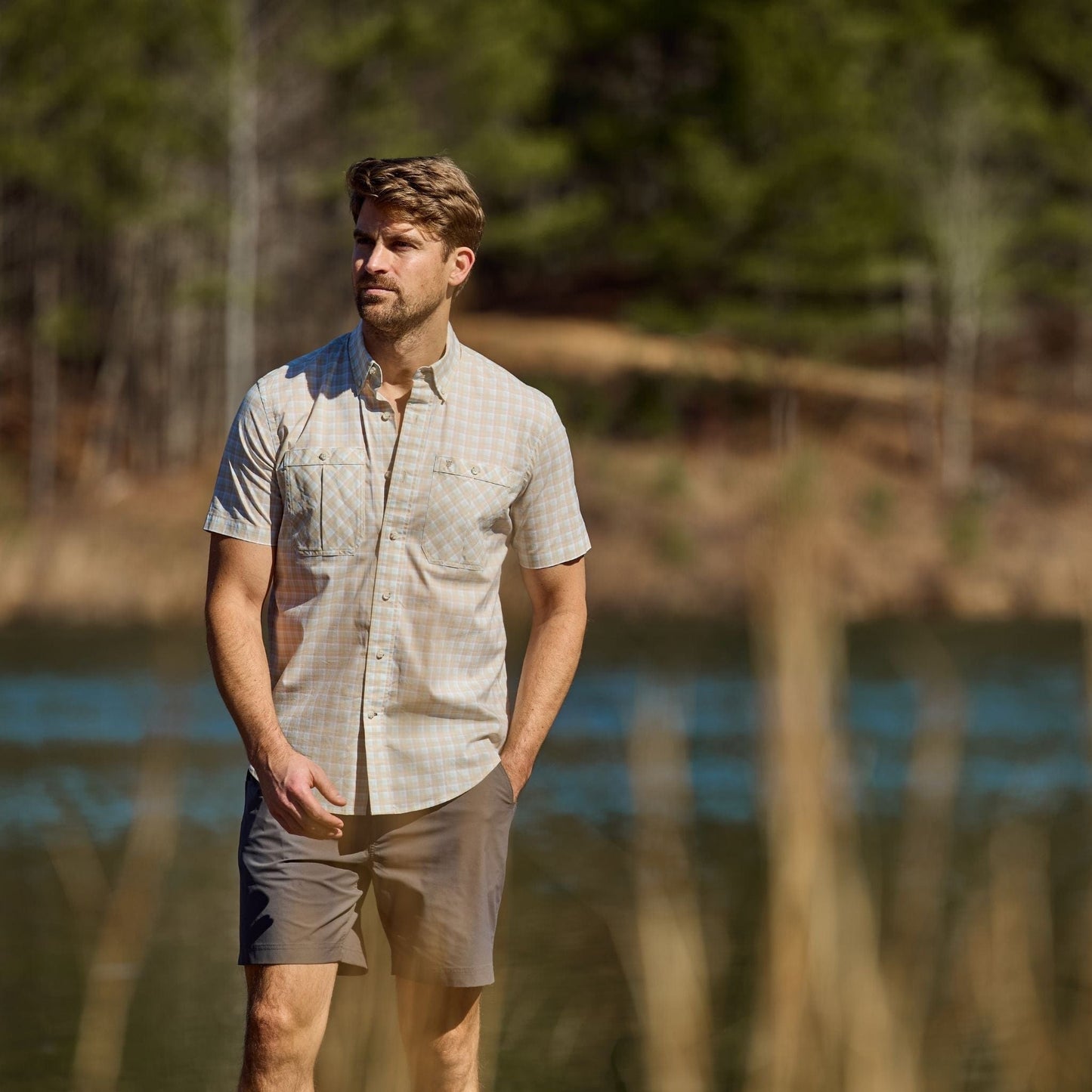 A man in a Tom Beckbe Tidewater short-sleeve shirt stands near a lake with trees in the background.