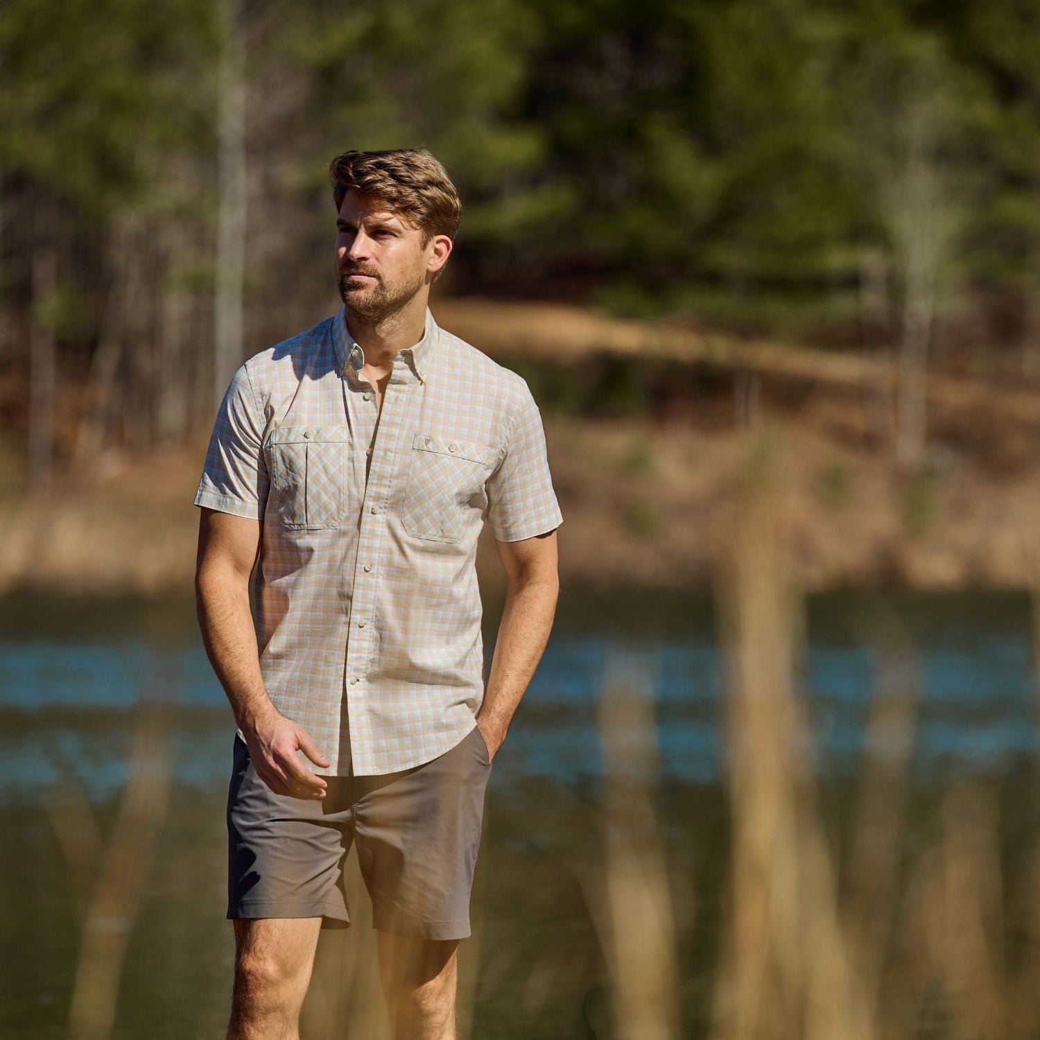 A man in a Tom Beckbe Tidewater short-sleeve shirt stands near a lake with trees in the background.
