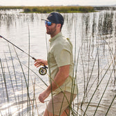 In a grassy water area, a person in a Tidewater Shirt by Tom Beckbe wears sunglasses and a cap, holding a fishing rod.