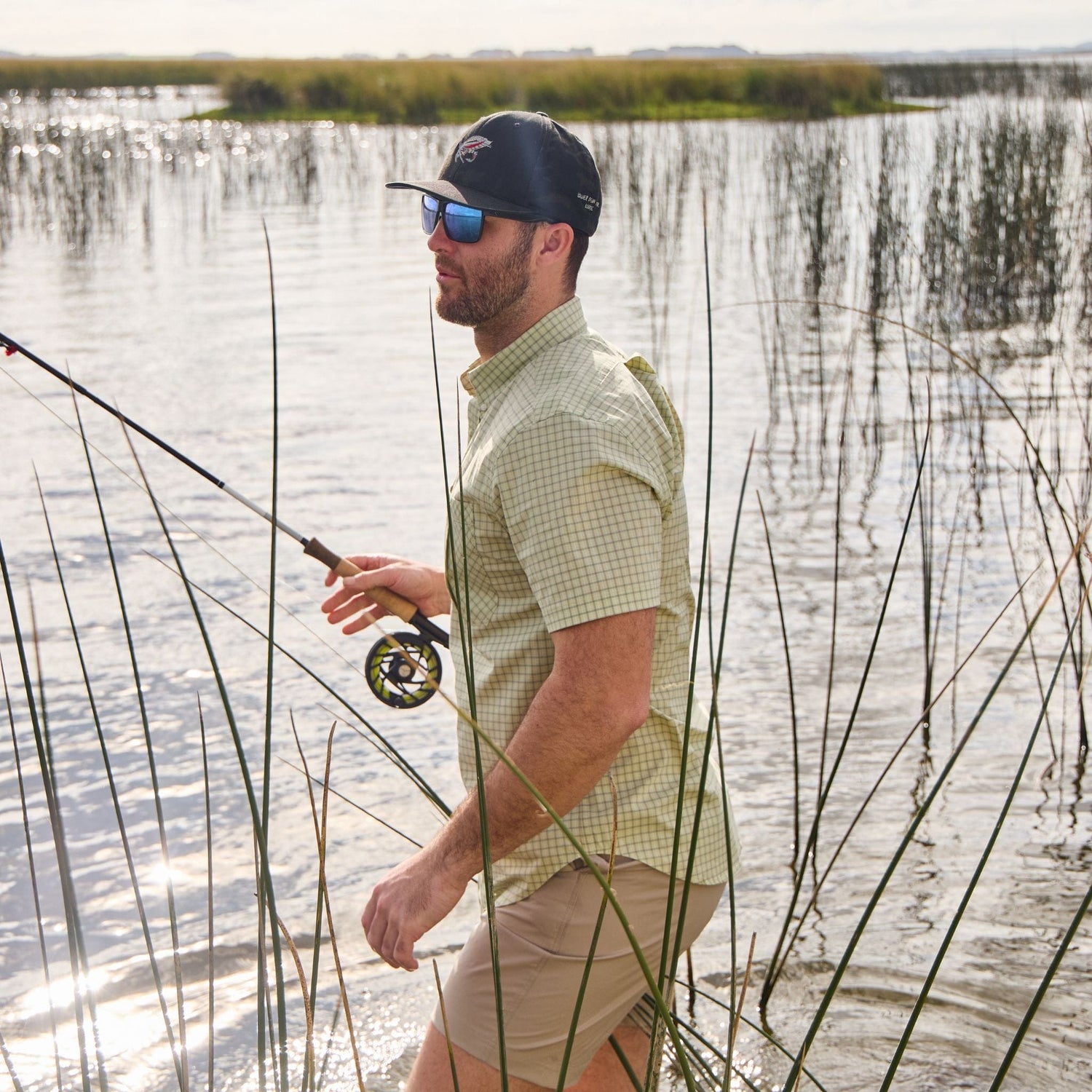 In a grassy water area, a person in a Tidewater Shirt by Tom Beckbe wears sunglasses and a cap, holding a fishing rod.
