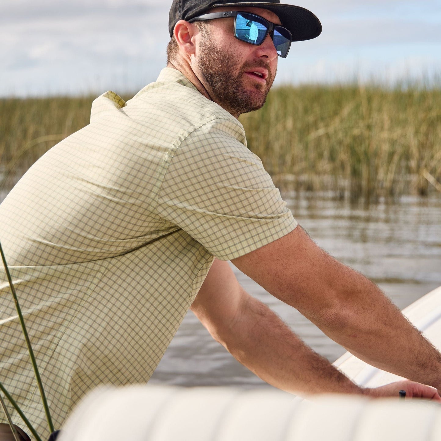 A man in sunglasses and a cap stands on a boat in a marsh, wearing the Tom Beckbe Tidewater Shirt.