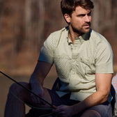 A man in a Tom Beckbe Tidewater Shirt holds a rod, sitting outdoors against a blurred forest backdrop.