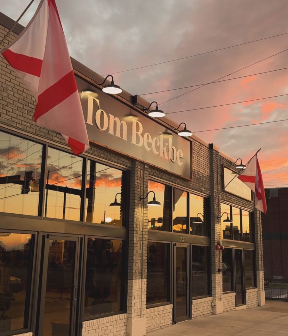 Building exterior with Tom Beckbe sign, displaying red and white flags, under a colorful sunset sky.