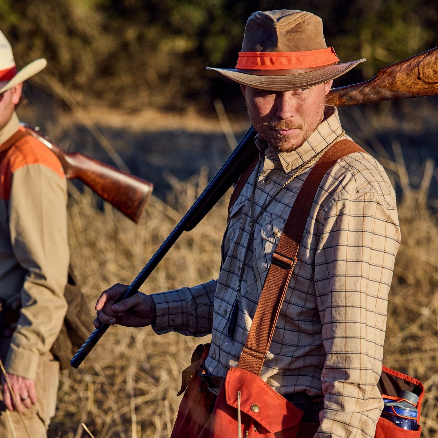 Two hunters with shotguns; one in focus dons a Tom Beckbe Field Hat Band and a nylon blaze orange vest.