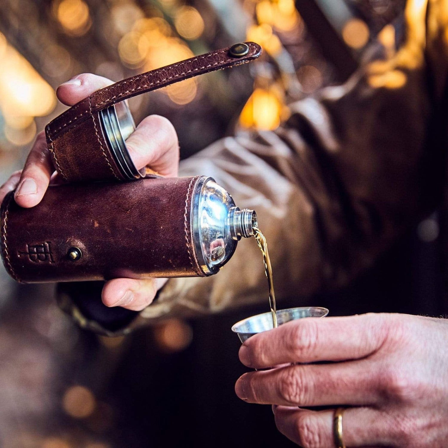 Man pours bourbon from a stainless bottle flask wrapped in leather into a stainless toasting cup.