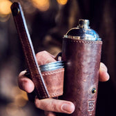 Man holds in his hand a stainless bottle flask wrapped in oak leather with embossed TB logo.