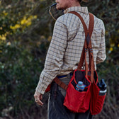 Person outdoors in Tom Beckbe Strap Vest, checkered shirt, hat, and red bag carrying bottles, sipping through a straw.