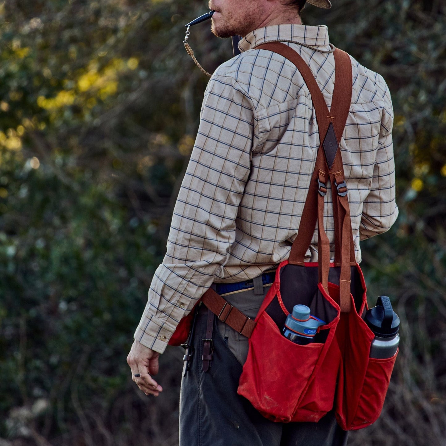 Person outdoors in Tom Beckbe Strap Vest, checkered shirt, hat, and red bag carrying bottles, sipping through a straw.