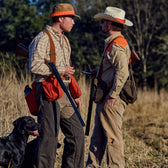 Two men in hunting gear with rifles and a black dog stand in a field; one wears a Tom Beckbe Strap Vest.