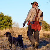 Man in Tom Beckbe Strap Vest with two black dogs walks through a field, carrying a hunting bag and wearing a brown hat.