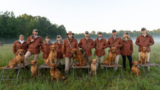 A group of nine people in matching brown jackets stand with six dogs in a grassy field with trees in the background.