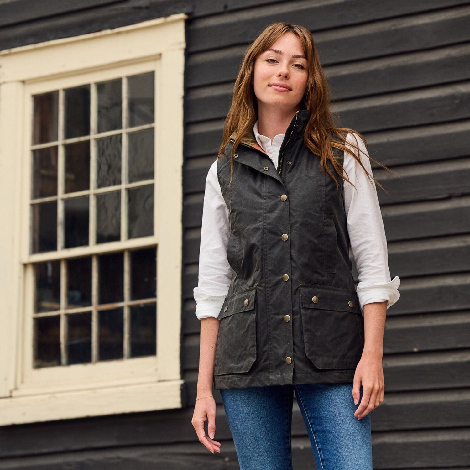 Person posing outdoors by a wooden wall, wearing a white shirt, Tom Beckbe Womens Fairmont Vest, and blue jeans.