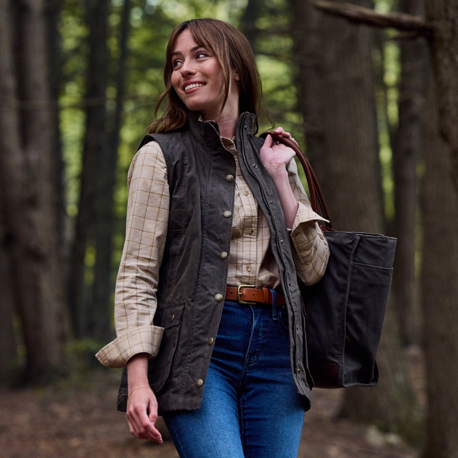Woman in a forest wearing a plaid shirt, jeans, Womens Fairmont Vest by Tom Beckbe, holding a tote bag and smiling.