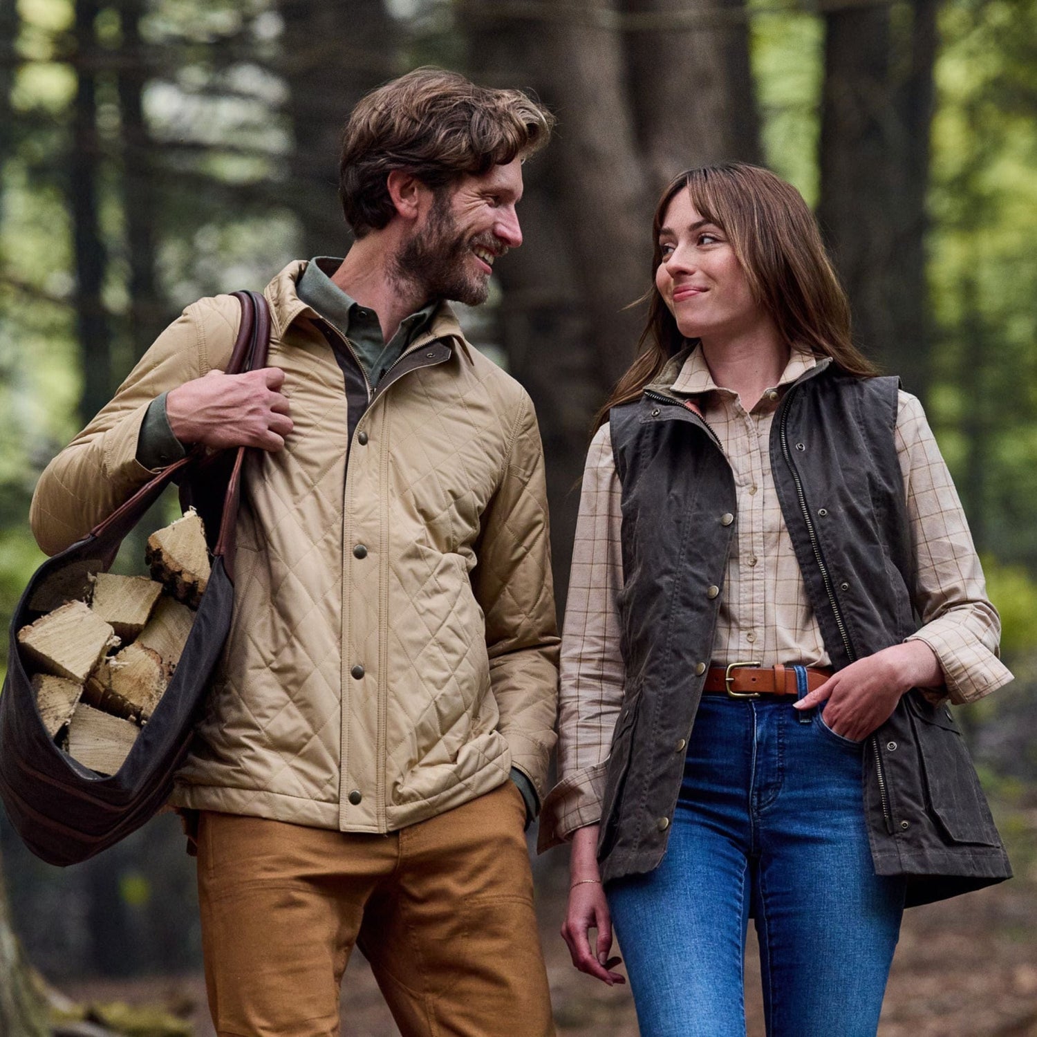 Two people stroll through the forest, one with firewood, both smiling in Tom Beckbes Womens Fairmont Vests.