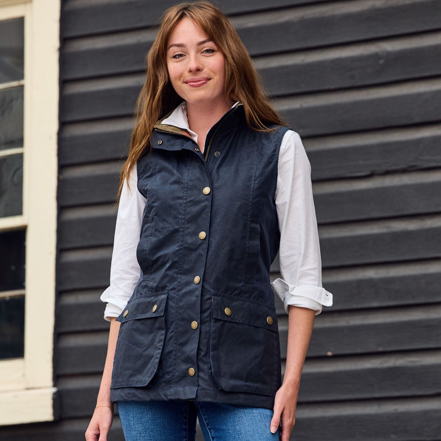 A person in a Tom Beckbe Womens Fairmont Vest over a white shirt stands by a dark wooden wall with a window.