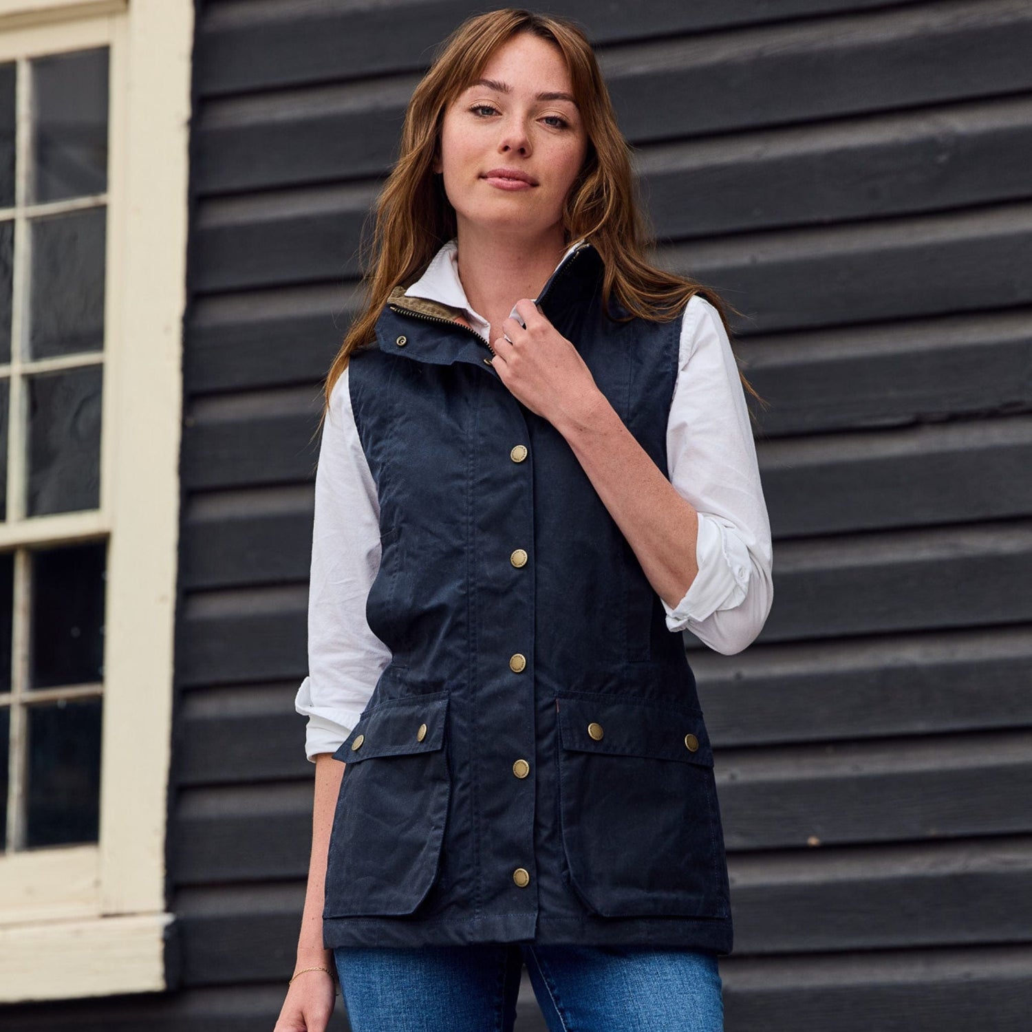 Person in a Tom Beckbe Womens Fairmont Vest over a white shirt, standing before a wooden building with a window.