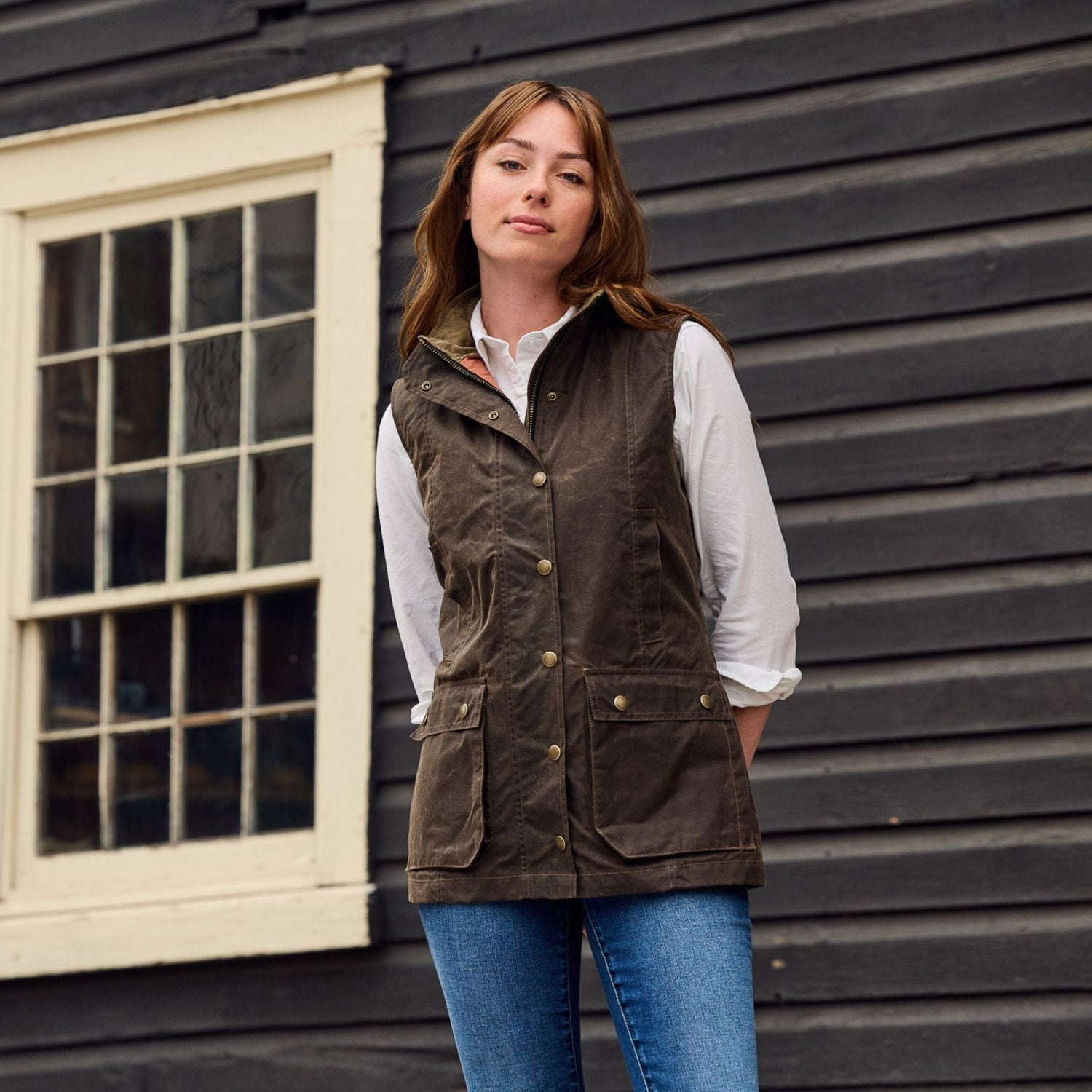 A person in a Tom Beckbe Fairmont Vest and blue jeans stands outside a black wooden building with white window frames.